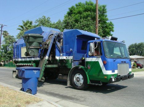 A large commercial rubbish collection truck parked on a suburban street with a predominantly blue and green color scheme. The truck features a front cab with a silver front grille and side mirrors, and an open-top hopper section in the back designed for waste collection, with a hydraulic lifting mechanism clearly visible. A green waste bin is positioned adjacent to the truck, ready for loading. The scene includes a paved road with a concrete curb, lush green trees, and residential houses visible in the background, indicating a typical local neighbourhood environment. The sky is clear and bright, suggesting daytime, and the overall setting aligns with waste management and rubbish removal services in Wanstead or nearby areas.
