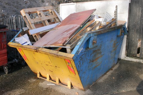 A large metal skip painted in yellow and blue, positioned on a concrete surface outdoors, partially filled with various discarded items including wooden pallets, metal sheets, and miscellaneous debris. The wooden pallets are stacked irregularly at the top of the skip, with some pieces leaning outward, while the metal sheets have a reddish-brown surface and are positioned among other waste materials. The background features a white wall and a section of a black wooden fence, indicating a residential or commercial property boundary. The environment appears to be an outdoor area in Wanstead, with natural daylight illuminating the scene, and the waste materials suggest a clearance or rubbish removal operation typical of services provided by Wanstead House Clearance.