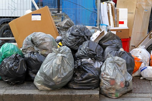 A collection of tightly packed black and grey plastic rubbish bags filled with waste material, arranged in a pile on a paved pavement area, with some bags leaning against a curb. The bags vary slightly in size and are situated outdoors, with a background that includes a blue container, cardboard boxes, and other miscellaneous waste items, suggesting a rubbish removal task in a residential or commercial area near Wanstead. The scene is well-lit, capturing the texture and creases of the plastic bags, illustrating typical waste awaiting collection or transportation, consistent with services offered by Wanstead House Clearance in the local area.