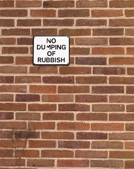 A rectangular metal sign mounted on a red brick wall, displaying the message 'NO DUMPING OF RUBBISH' in black capital letters on a white background. The sign is positioned centrally in the upper left