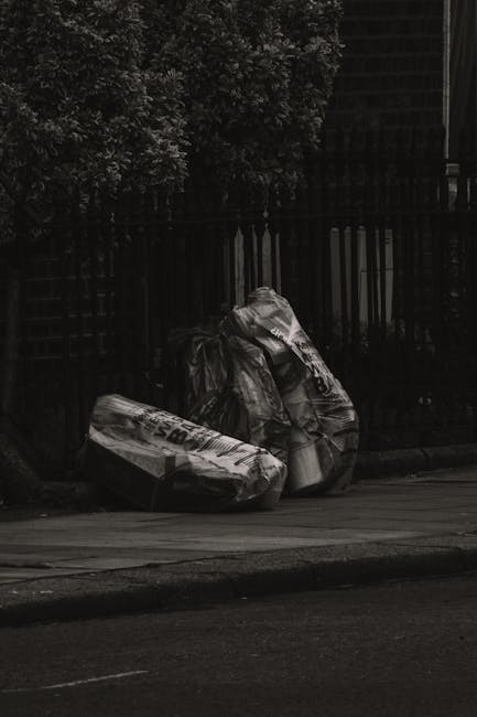 Two large black plastic garbage bags filled with waste materials are positioned on the pavement next to a black metal fence with vertical bars. The bags appear to be tightly packed and tied at the top