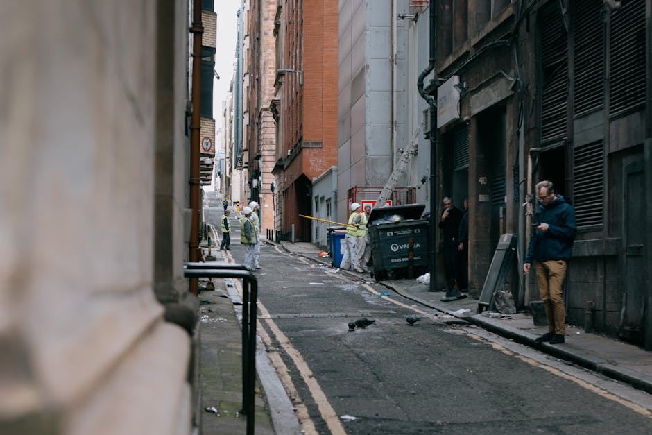 A narrow, paved alleyway leading into a private area, flanked by green metal fences on both sides with vertical bars and a wooden gate on the right. The scene features a tall, grey utility pole positi