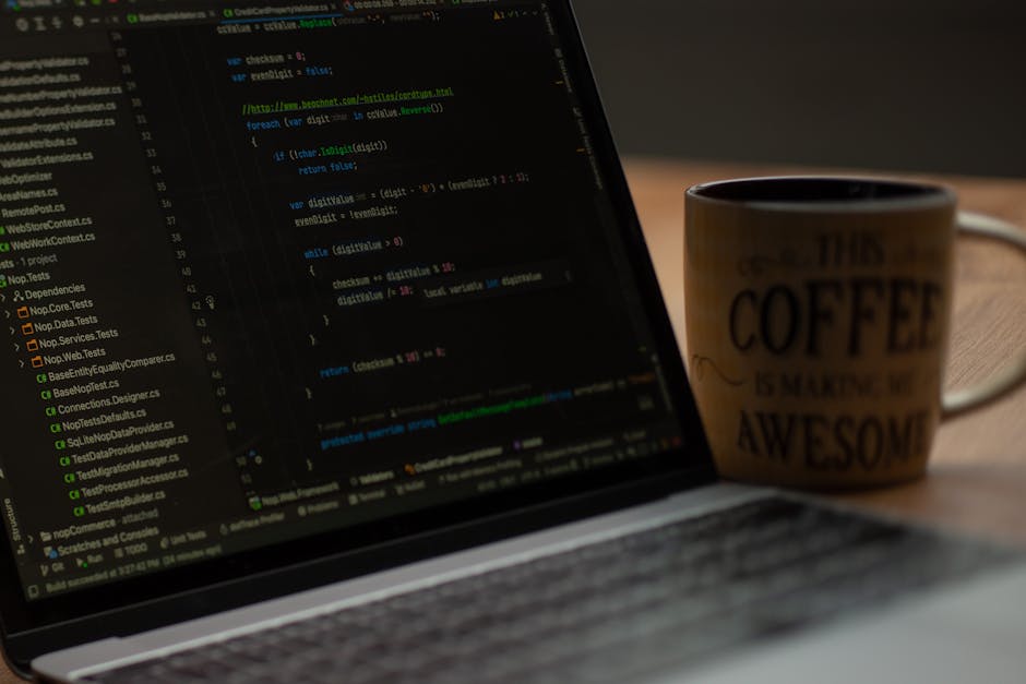 A person with a wristwatch on their left wrist is seated at a wooden desk, using a silver laptop with a black keyboard. The laptop screen displays lines of code or text in a dark-themed editor, with t